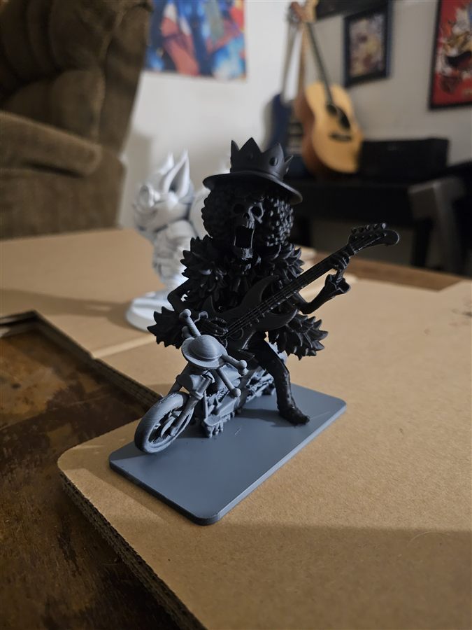Black 3D-printed figure of Brook from One Piece playing guitar, next to a gray motorcycle with a straw hat on the handlebars