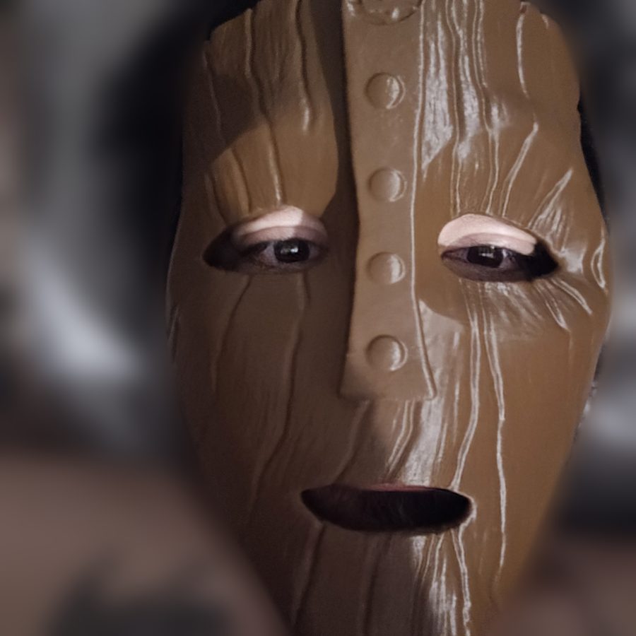 Person wearing a brown 3D-printed wooden-looking mask with a studded center strip, with the background blurred