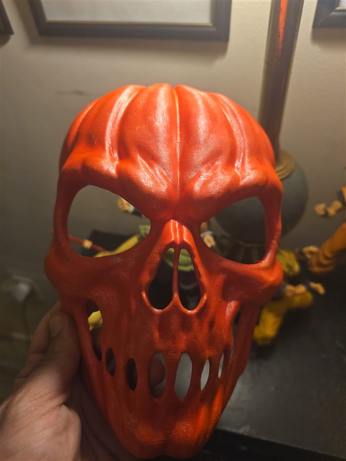 Hand holding a red-orange 3D-printed mask shaped like a pumpkin skull, with a ribbed pumpkin top, open eye holes, nose holes and slotted teeth