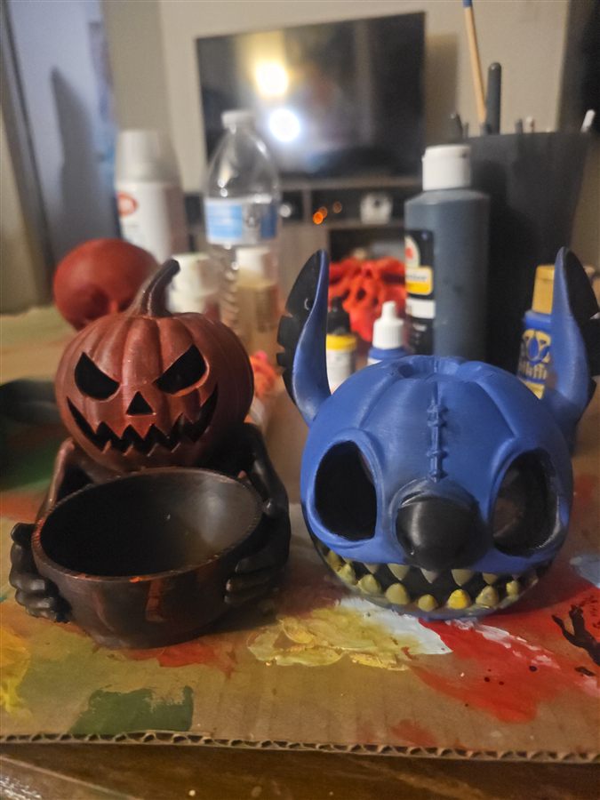 Red-orange 3D-printed jack-o'-lantern with an angry carved face, sitting on a dark skeleton body that holds a large bowl, next to a blue Stitch head with sharp teeth