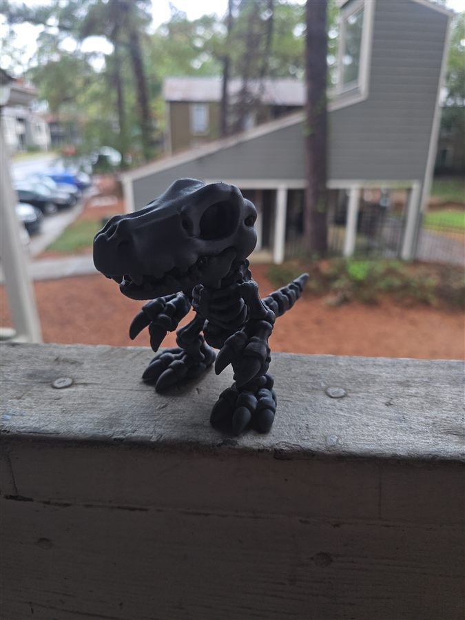 Small black 3D-printed Skeleton Agumon from Digimon standing on a wooden railing outdoors