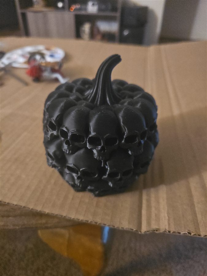 Black 3D-printed pumpkin made of stacked skulls with a ribbed stem, sitting on a cardboard box