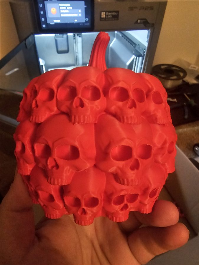Hand holding a red 3D-printed pumpkin made entirely of stacked skulls, with the Bambu Lab P2S printer visible behind it
