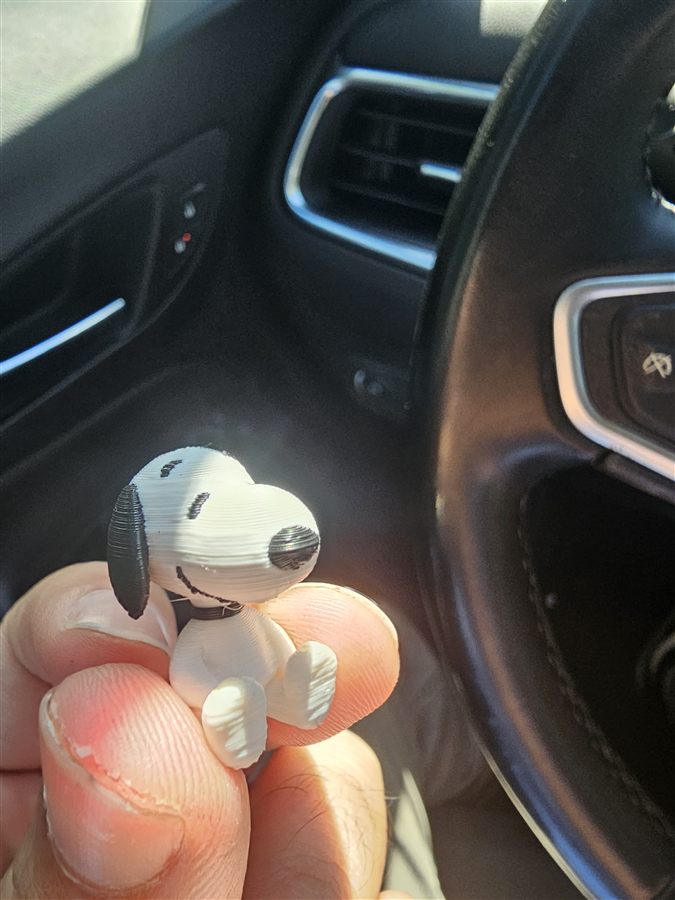 Tiny white and black 3D-printed Snoopy figure held in front of a car steering wheel
