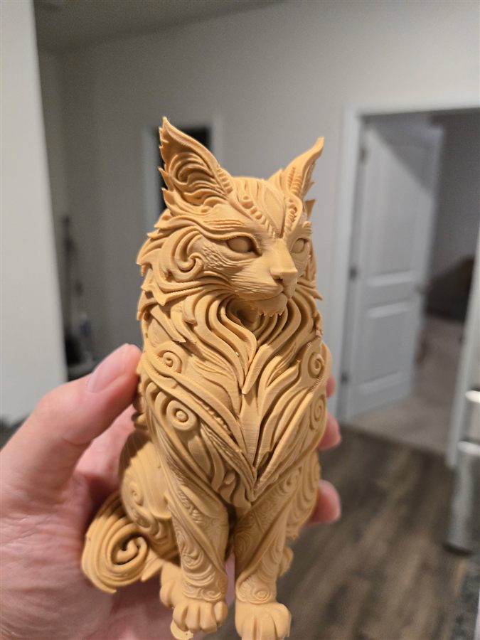 Hand holding a tan 3D-printed long-haired cat sitting upright, with fur carved into swirling ornate patterns