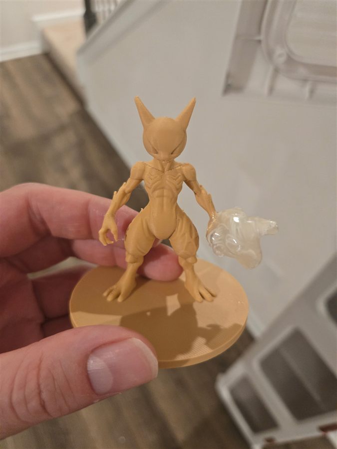 Hand holding a small tan 3D-printed figure with pointed ears and defined muscles, standing on a round base with a clear translucent blob in one hand