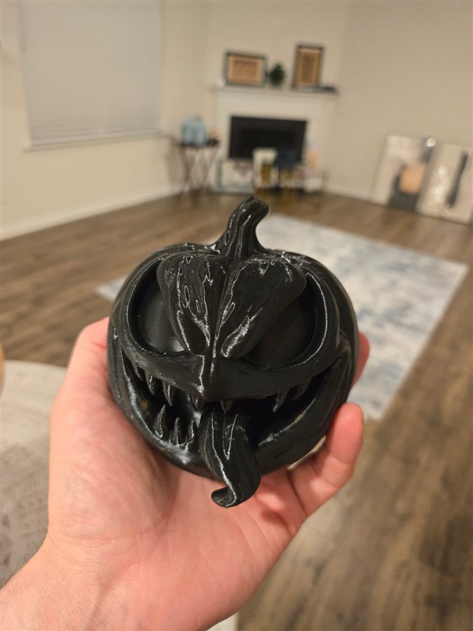 Hand holding a black 3D-printed jack-o'-lantern with angry eyes, sharp fangs, and a long tongue hanging out
