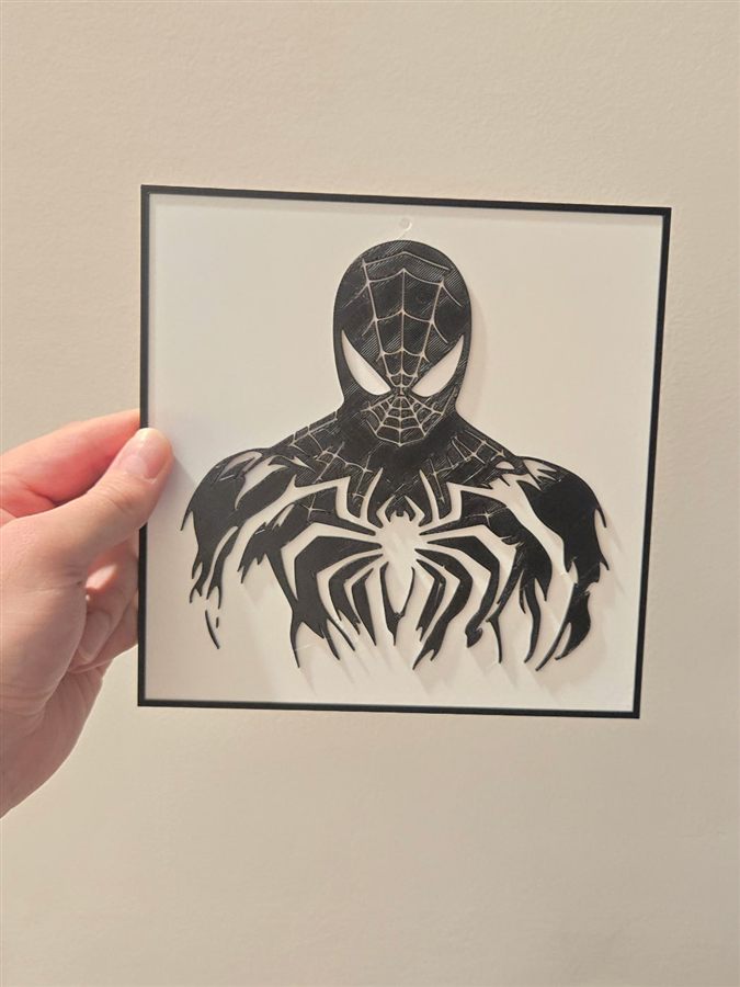 Hand holding a square black-and-white 3D-printed wall plaque of Spider-Man's upper body in a black suit with a white spider emblem, in a thin black frame